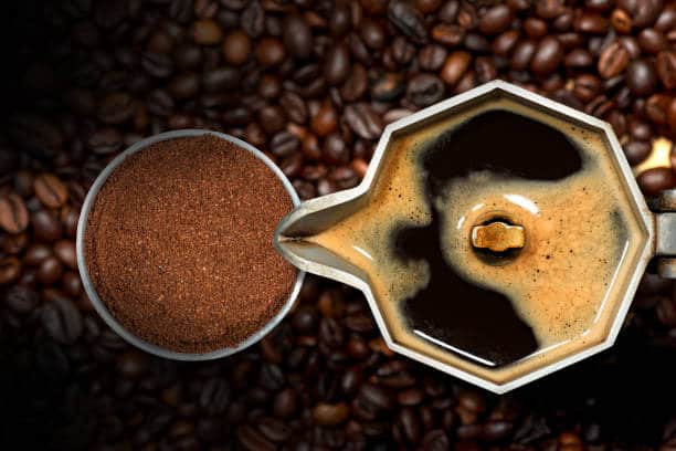 Close-up of an open Italian coffee maker with ground coffee in a metal filter, on many roasted coffee beans, top view