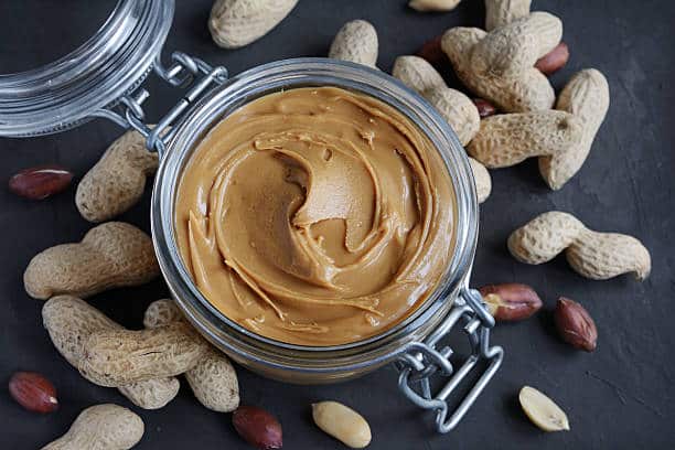 Peanut butter in a glass jar and peanuts on a black slate background