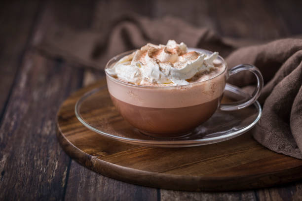 Hot chocolate with whipped cream in a glass cup. The cup is on a glass saucer, on a wooden background. The chocolate is sprinkled with cocoa powder. There is a brown llinen cloth in the background.