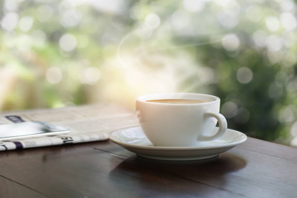 A cup of coffee on teak wood table with blurred mobile phone on newspaper, garden bokeh background.