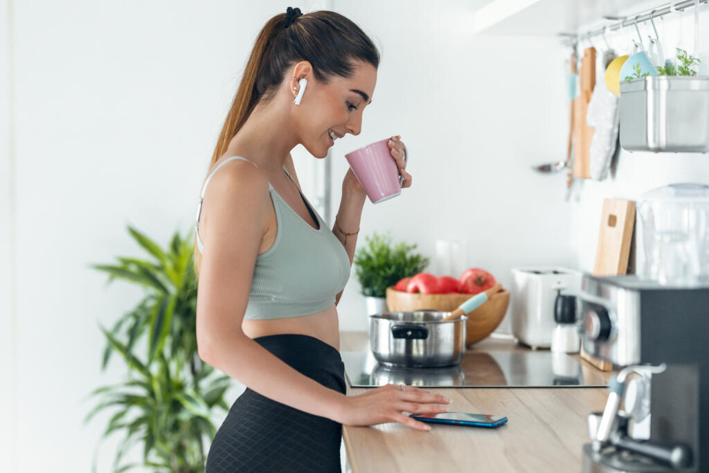 Shot of sporty woman listening to music with her smartphone whiledrinking coffee in the kitchen at home.