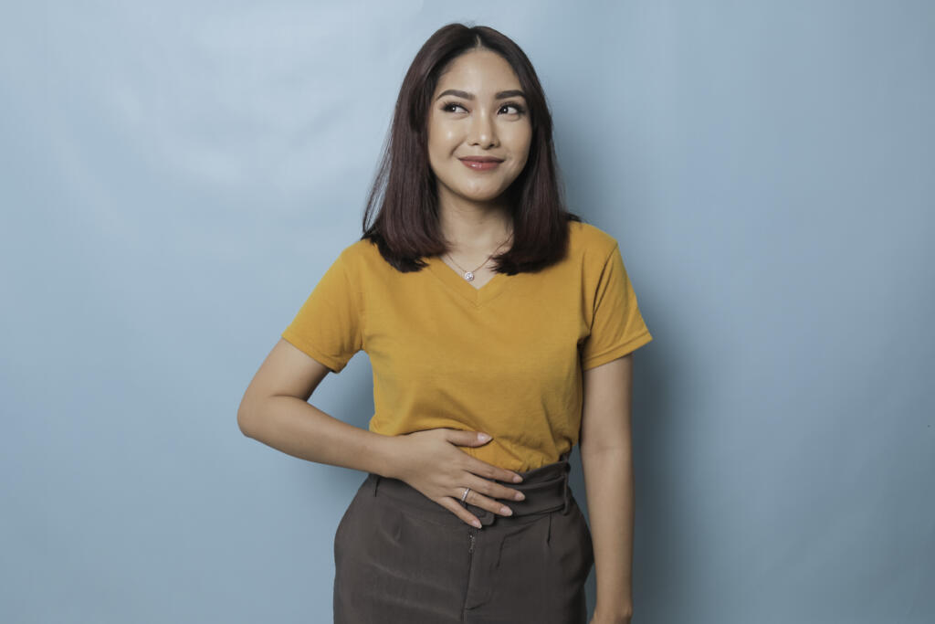 Pleased cheerful Asian woman keeps hand on belly, feels full after a delicious dinner dressed casually and stands thoughtful against blue background.