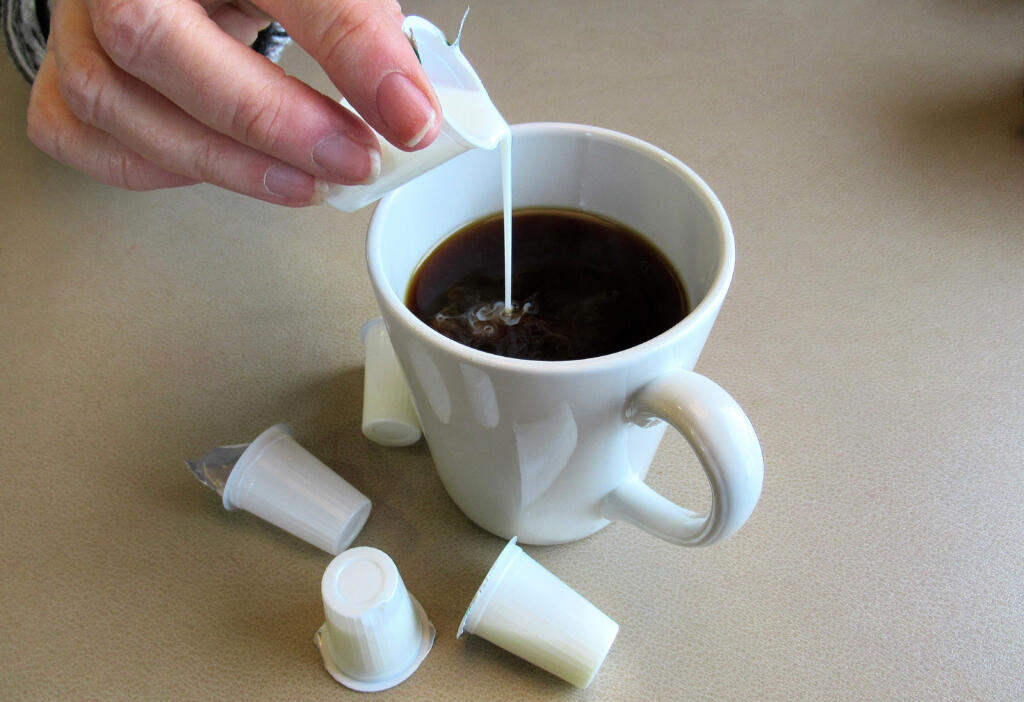 A close up shot of milk, being poured into a cup of black coffee, from a small, disposable, plastic container..